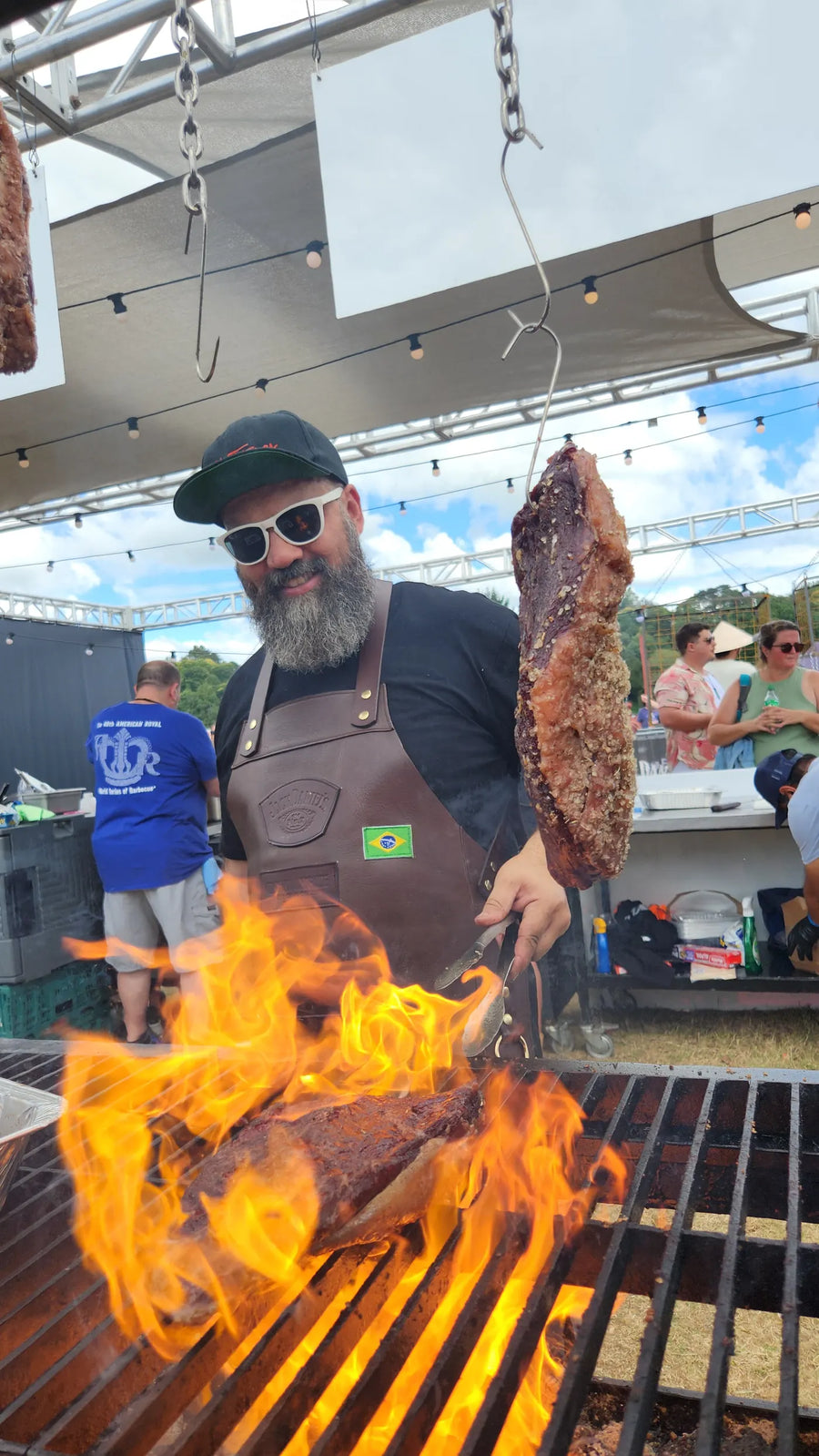 Adriano Andrade lifting a rack of beef ribs off a live fire at an outdoor cook, flames coming up through the grate